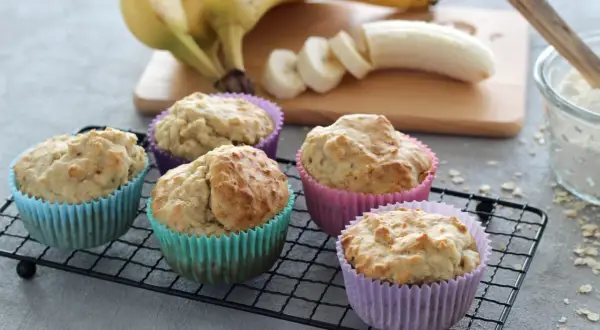 Fünf leckere Muffins ohne Zucker in bunten Förmchen, daneben Bananen und eine Schüssel Mehl.
