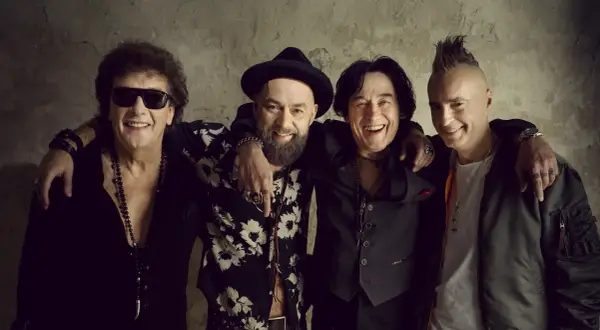 Czterech mężczyzn z zespołu Lady Pank, w tym jeden w okularach przeciwsłonecznych i z burzą loków, drugi w kapeluszu i z brodą, trzeci z charakterystyczną fryzurą i czwarty w kurtce bomberce, pozuje do zdjęcia.