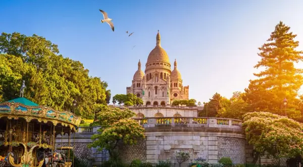 Sacre Coeur reser sig majestätiskt mot en klarblå himmel, med en karusell i förgrunden och en mås som svävar ovanför.