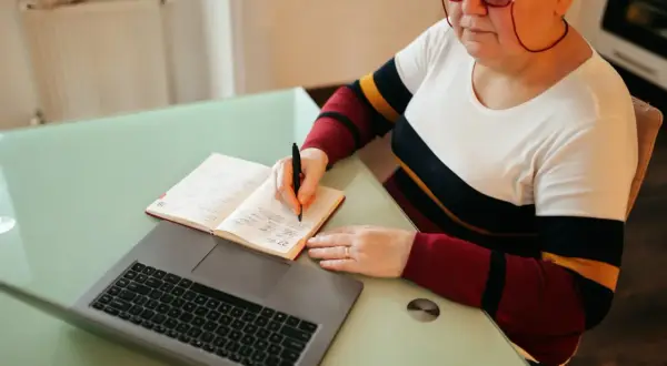 Kobieta w okularach pisze w notatniku obok laptopa.