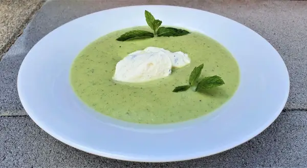 Velouté de courgette onctueux, garni d'une quenelle de fromage frais et de feuilles de menthe, servi dans une assiette blanche.