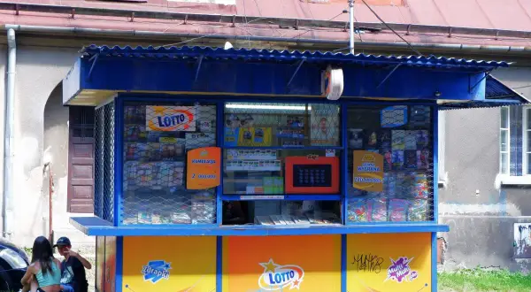 Kiosk Lotto z ofertą zdrapek. Czy warto kupować zdrapki na stacjach benzynowych czy lepiej w kolekturze? Dwoje młodych ludzi siedzi obok.