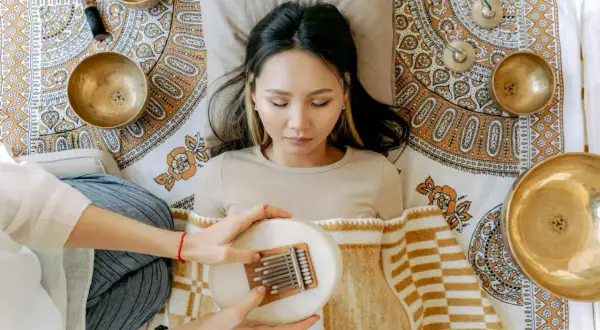 Eine Frau liegt entspannt, während eine andere Person ihr eine Kalimba über dem Bauch hält. Klangschalen und ein Stößel sind um sie herum arrangiert, was auf eine Klang Meditation hindeutet.