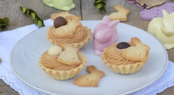 Wielkanocne babeczki z mascarpone, ozdobione ciasteczkami w kształcie króliczków i czekoladowymi jajeczkami, na talerzu wśród ceramicznych zajączków.