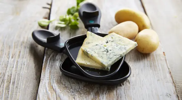 Tranches de bleu de Gex prêtes pour la raclette, avec des pommes de terre et de la verdure sur fond de bois.