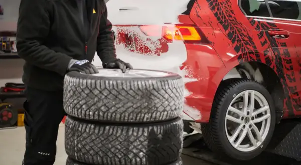 Person mit Handschuhen stapelt Winterreifen neben einem roten Auto mit Schneespritzern und Reifenprofil-Aufklebern.