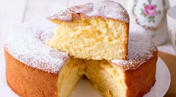 En fluffig vegansk sockerkaka, pudrad med florsocker. En bit är upplagd ovanpå resten av kakan.