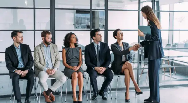 Un grupo de personas espera en fila para una entrevista de trabajo. Una mujer sonríe mientras le da la mano a otra mujer, quizás sintiendo que no me adapto a mi nuevo trabajo.