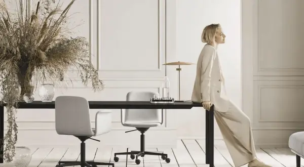 Eine Frau in einem beigen Anzug sitzt auf einem schwarzen Tisch. Der Raum ist in neutralen Farben gehalten und mit Trockenblumen und Glasvasen dekoriert.
