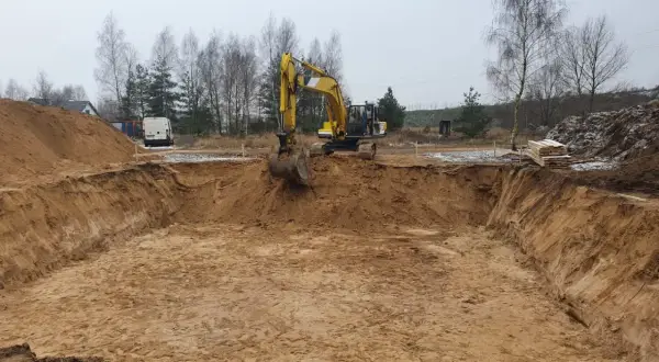 Koparka pracuje przy wykopy pod fundamenty. Widać głęboki dół, usypiska ziemi i materiały budowlane.