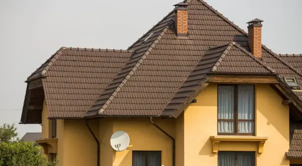 Dach naczółkowy konstrukcja z brązowej dachówki, z ceglanym kominem i anteną satelitarną na żółtej ścianie domu.