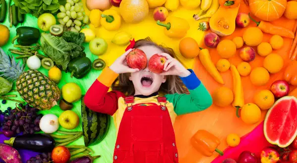 Enfant joyeux avec des pommes devant les yeux, entouré de fruits et légumes colorés. Une belle scène pour une activité manuelle semaine du goût.