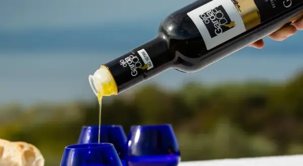 Versando olio Garda DOP in bicchierini blu, con pane fresco sullo sfondo.