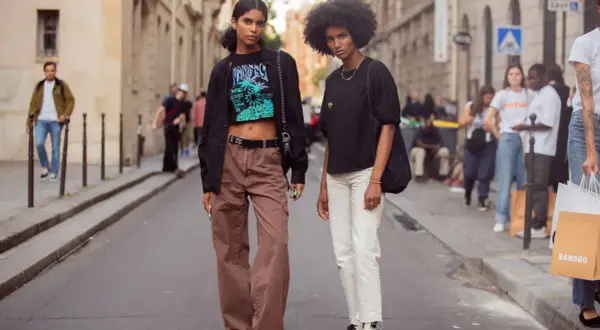 Zwei Frauen in lässiger Kleidung, eine mit brauner Cargohose und Crop-Top, die andere mit weißer Hose und schwarzem Shirt, stehen auf einer Straße.