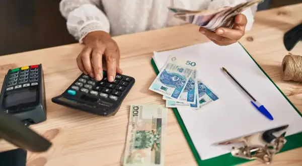 Kobieta liczy pieniądze na kalkulatorze, przygotowując się do rozliczeń związanych z samozatrudnieniem. Na stole leżą polskie banknoty.