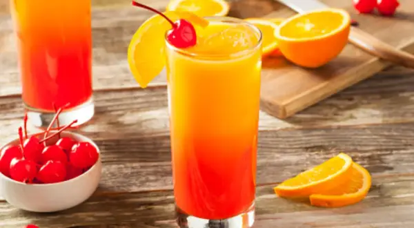 Cóctel refrescante con capas de granadina y naranja, adornado con una cereza y rodajas de naranja.