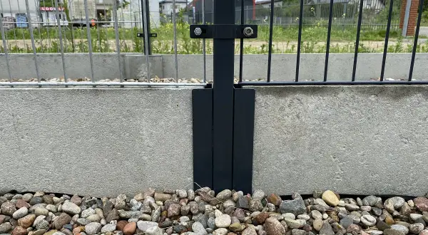 Metalowy łącznik słupka ogrodzenia do betonowej podmurówki. Kamienie ozdobne wokół.