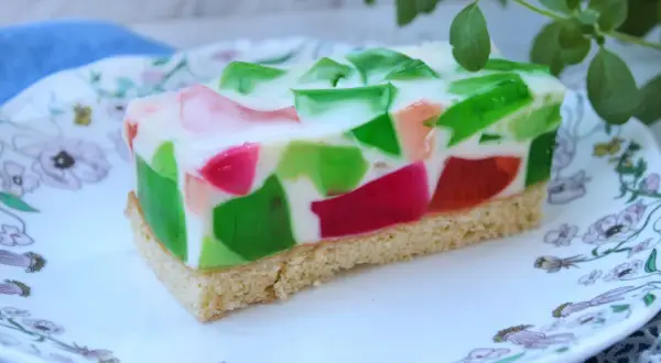 Kolorowe ciasto z galaretkami w kostkę, z zielonymi i różowymi kawałkami zatopionymi w białej masie, na spodzie biszkoptowym.