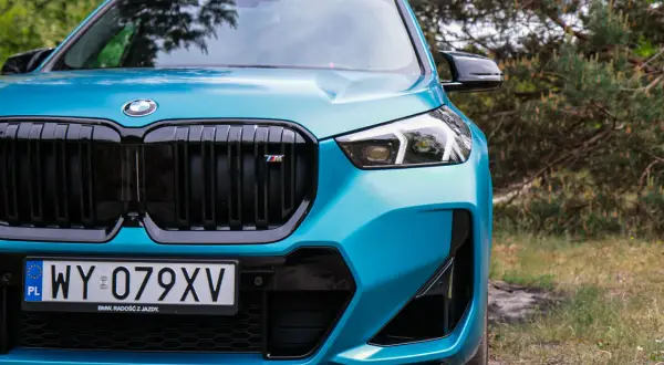 Turkusowy BMW X1 2024 z czarnym grillem i emblematem M, gotowy na przygodę w otoczeniu natury.