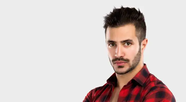 Un homme au regard intense, avec une barbe mouche bien taillée, porte une chemise à carreaux rouges et noirs.