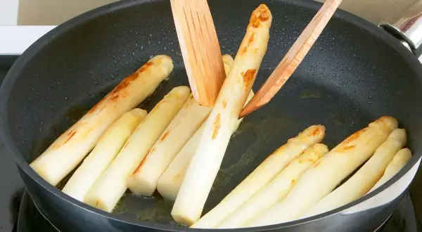 Weißer Spargel brät in einer Pfanne mit etwas Öl. Holzspieße wenden die Stangen, die leicht gebräunte Stellen zeigen.