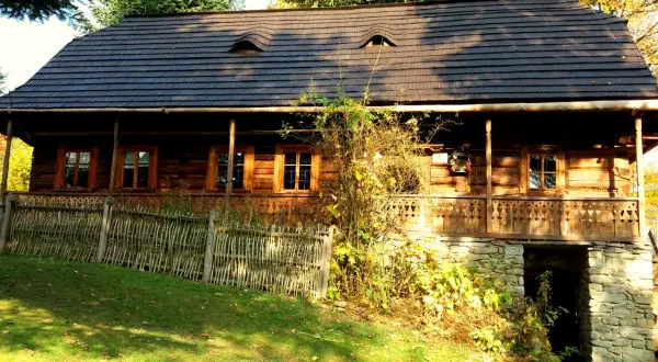 Drewniany dom z kamienną piwniczką i ozdobną werandą, otoczony zielenią i jesiennymi liśćmi.