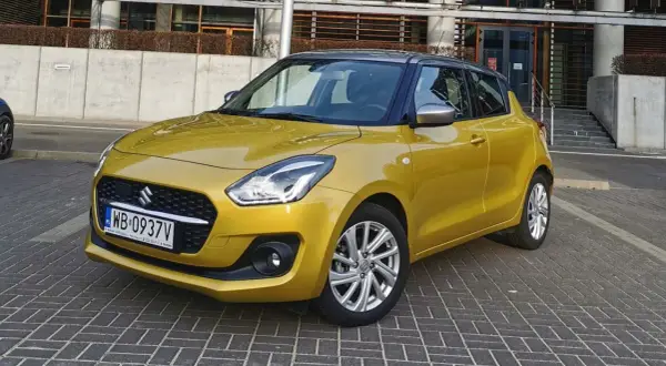 Złoty Suzuki Swift na tle nowoczesnego budynku. Wiele pozytywnych suzuki swift opinie potwierdza jego miejski charakter.