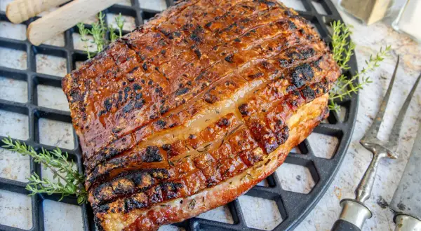 Perfekt gegrillter Schweinebauch mit knuspriger Schwarte liegt auf einem Grillrost. Kräuter und Grillbesteck deuten auf ein köstliches Mahl hin.