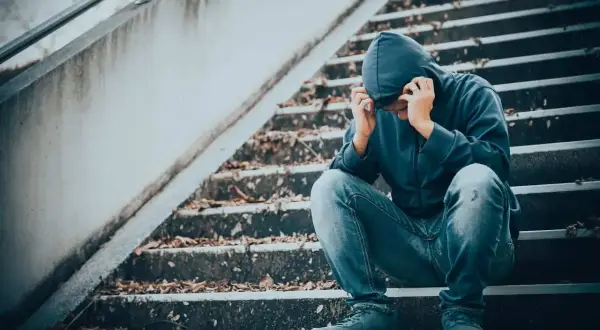 Młoda osoba w kapturze siedzi na schodach, pogrążona w rozpaczy. Obraz symbolizuje mechanizmy uzależnienia od narkotyków i walkę z nimi.