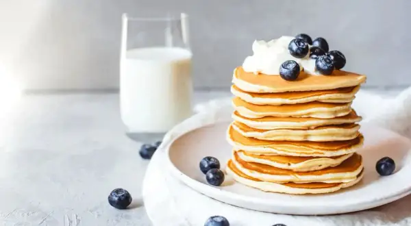 Ein Stapel goldbrauner Protein Pfannkuchen, garniert mit Sahne und Blaubeeren, daneben ein Glas Milch.