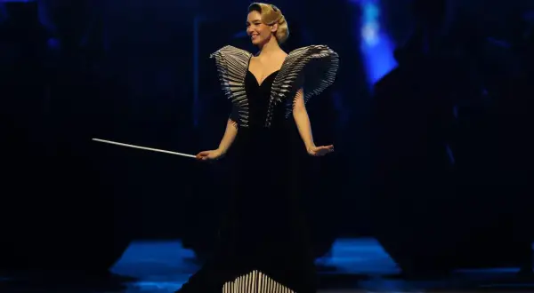 Matilda De Angelis in einem dramatischen schwarzen Kleid mit auffälligen Schulterdetails und einem Dirigentenstab.