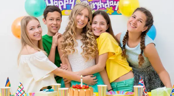 Uśmiechnięci nastolatkowie świętują urodziny przy stole pełnym balonów i tortu. W tle baner "Happy Birthday". Idealne konkursy zabawy na 50 urodziny!