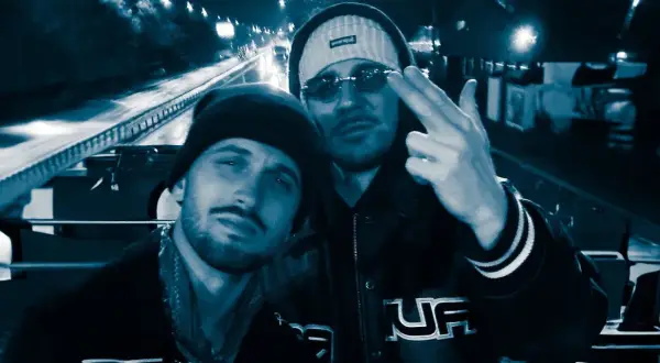 Dwóch mężczyzn w czapkach i okularach przeciwsłonecznych, jeden pokazuje gest ręką. W tle nocna droga, jakby pezet obejmuję noce.