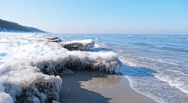 Lodowe sople zwisają z zamarzniętych kęp śniegu na plaży, gdy fale Bałtyku delikatnie obmywają brzeg. To widok z roku, kiedy zamarzł bałtyk rok.