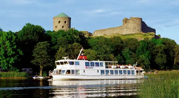 En vit båt från Strömma Boat Tours glider fram på vattnet, förbi grönskande träd och en gammal borg.