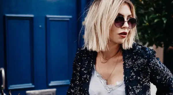 Frau mit lässiger hipster frisur, Sonnenbrille und Paillettenjacke vor blauer Tür.