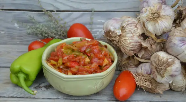 Un bol de piperade basque aux poivrons verts, tomates et oignons, avec des gousses d'ail et des herbes sur fond de bois.