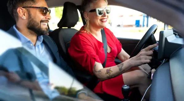 Kobieta w czerwonej sukience i okularach prowadzi auto, obok siedzi mężczyzna w okularach. Qarson w tle.