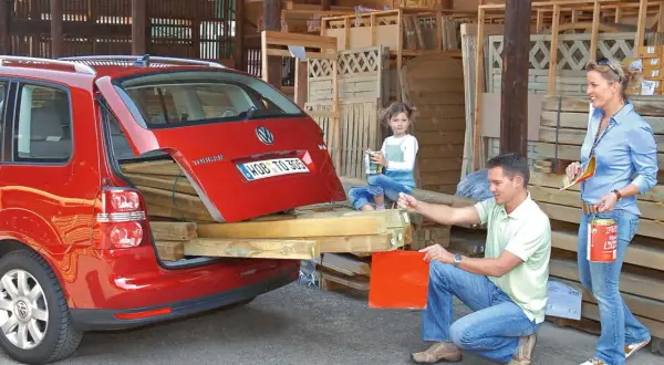 Familie belädt roten Kombi mit Holzbrettern. Die Heckklappe ist offen, die **Ladung nach hinten** überstehend. Ein Mann und eine Frau packen an, ein Kind schaut zu.