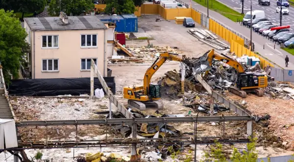 Dwa koparki pracują przy rozbiórce budynku. W tle widać ruchliwą ulicę i zaparkowane samochody.