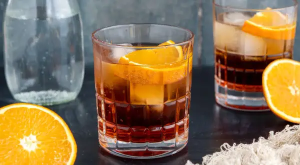 Ein erfrischender Americano Cocktail mit Eis und Orangenscheiben, daneben eine Karaffe Wasser und frische Orangen.