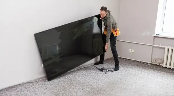 Mężczyzna w pasie narzędziowym trzyma duży, czarny telewizor przy ścianie.