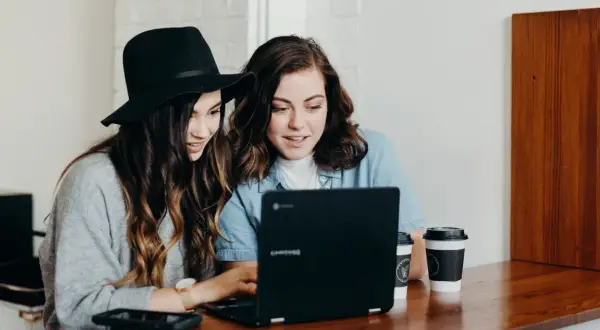 Två unga kvinnor diskuterar sin skuldkvot och bolån framför en laptop, med kaffekoppar på bordet.