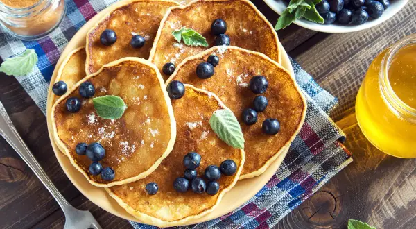 En hög med nyttiga pannkakor toppade med blåbär och pudersocker. Bredvid står en burk honung och en skål med mer blåbär.