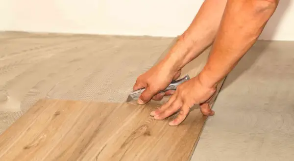 Hands using a utility knife to score and cut a vinyl plank flooring piece, demonstrating how to cut vinyl plank flooring for a perfect fit.