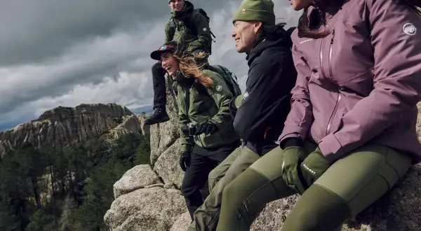 Grupa przyjaciół w zielonych i fioletowych kurtkach softshell i hardshell siedzi na skale, podziwiając górski krajobraz.