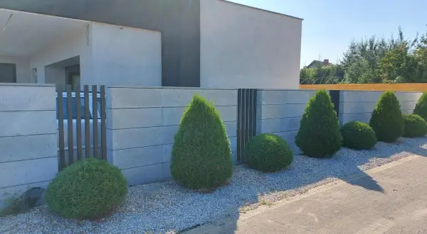 Nowoczesny betonowy płot z ozdobnymi krzewami i bramą.