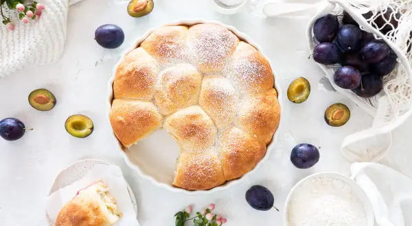 Ein goldbrauner Hefekuchen mit Puderzucker, umgeben von frischen Pflaumen. Perfekt für ein Buchty Rezept!