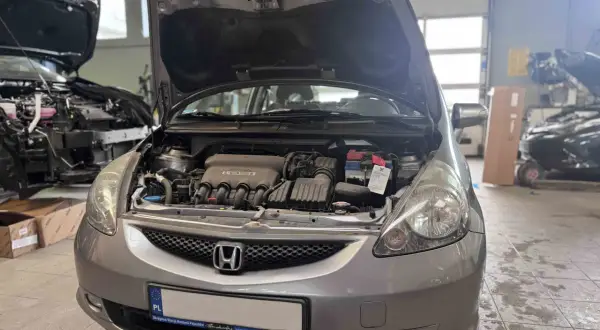 Srebrna Honda Jazz z otwartą maską, widoczny silnik i akumulator. Wymiana żarówki Honda Jazz w warsztacie.