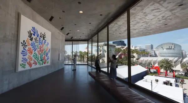 Inside a modern art museum, a colorful Matisse cutout hangs on a concrete wall. Visitors admire the art, enjoying the view of the city skyline.